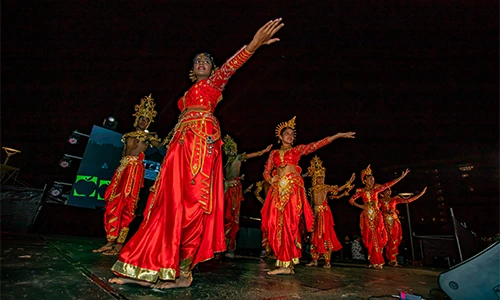 Group of dancers in red and gold traditional costumes performing on a dark stage under focused lighting.